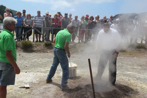 Köche heben in Furnas einen Topf Cozido aus einem dampfenden Loch im vulkanischen Boden, während Besucher zuschauen und fotografieren
