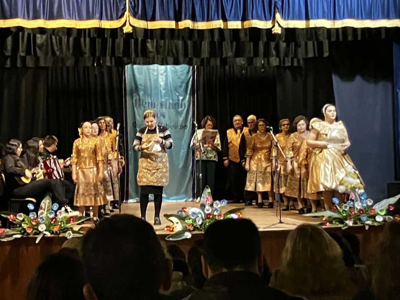 Eine Bailinho-Gruppe in goldenen Kostümen tritt auf der Bühne eines Gemeindesaals auf, daneben Musiker