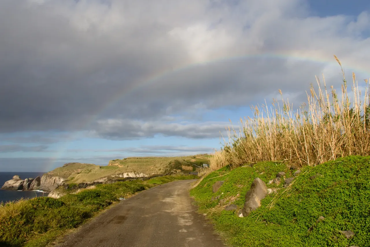 Rainbow over a coastal road and green cliffs on São Miguel, with sun breaking through grey rain clouds