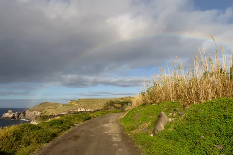 Arcoíris sobre una carretera costera y acantilados verdes en São Miguel, con el sol asomando entre nubes grises de lluvia