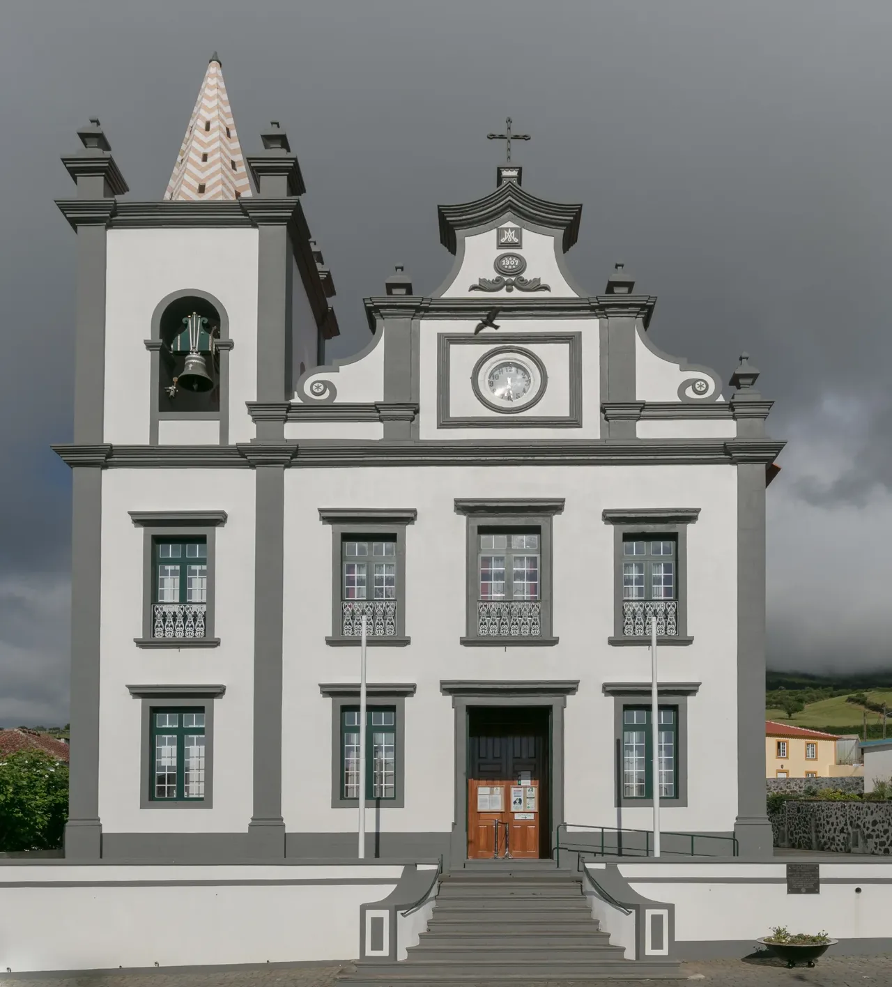 Die weiß-graue Fassade der Kirche von Serreta mit Uhr und Glockenturm unter dunklem Himmel